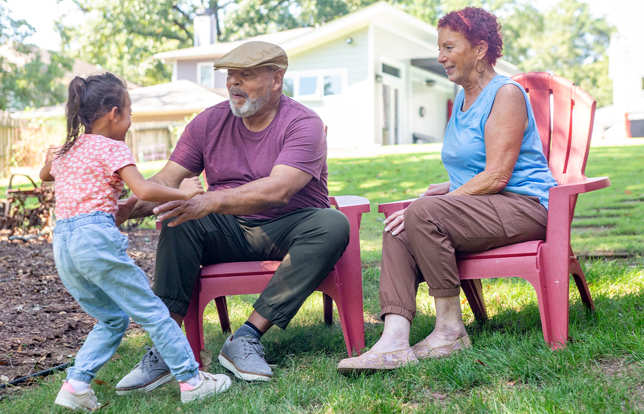 Man and woman sitting outside in chairs, while a young girl is walking towards the man with hands out and about to be picked up