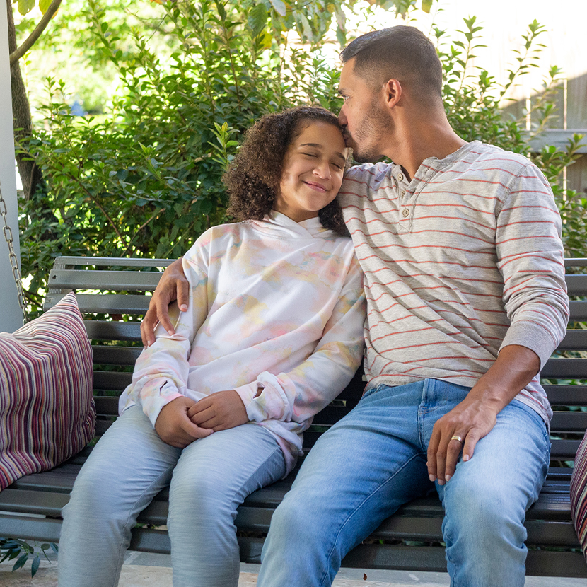 Man and adolescent female sitting on a swing outside, man kissing female on the forehead