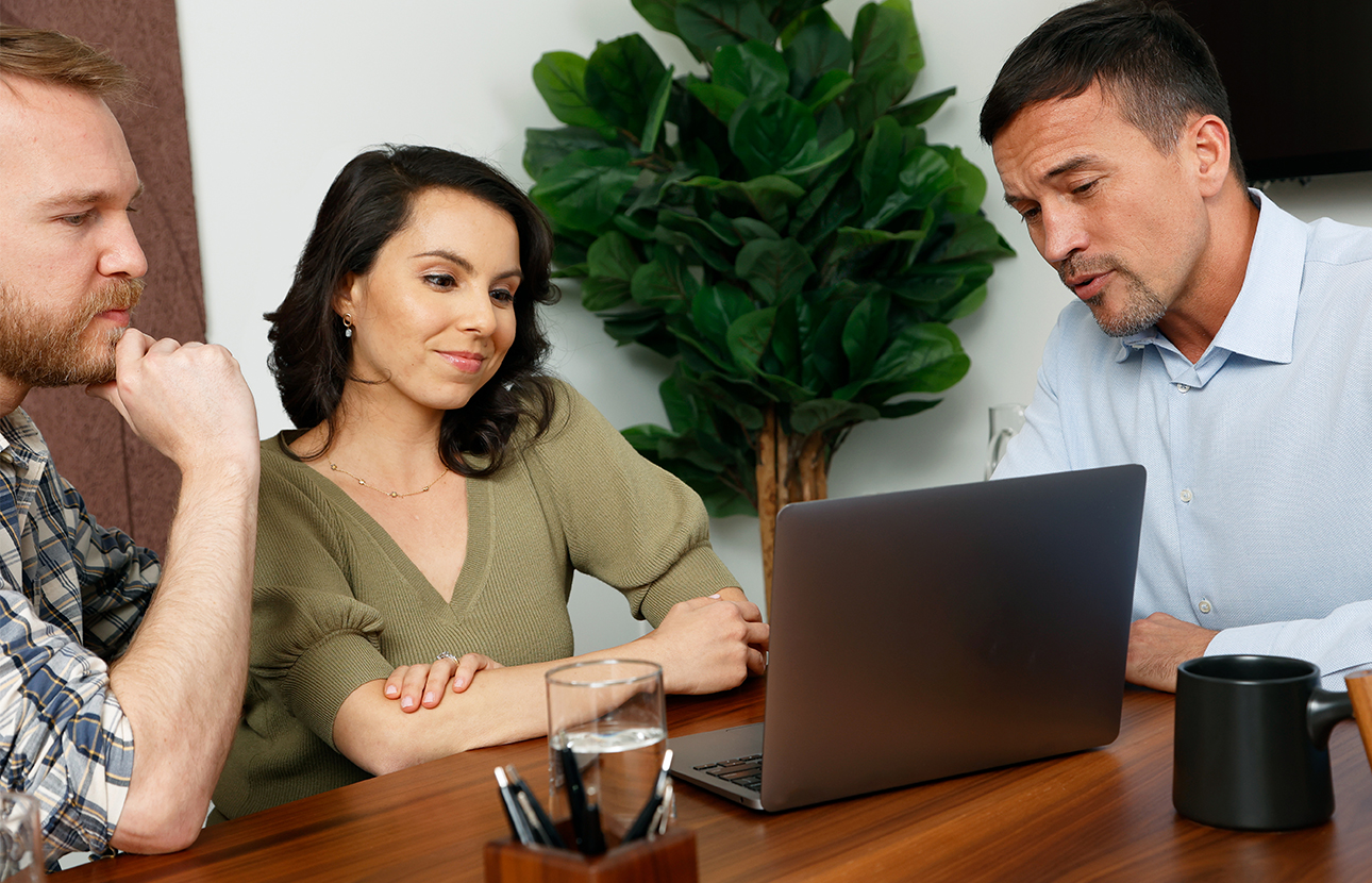 Two men and a woman sitting at a table, all looking at a laptop computer