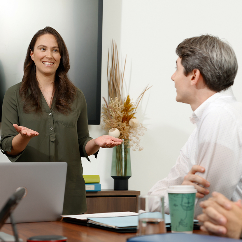 A woman and a man in an office setting, the woman standing and smile while the man is sitting at a table looking at her
