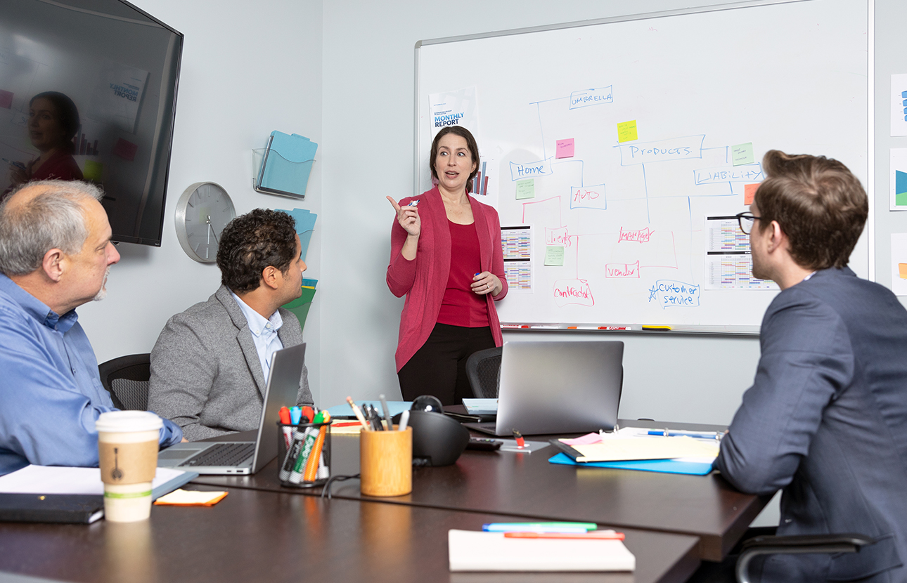 A woman and two men in an office setting, the men are sitting around a table and the woman is standing next to a white board with a marker in her hand