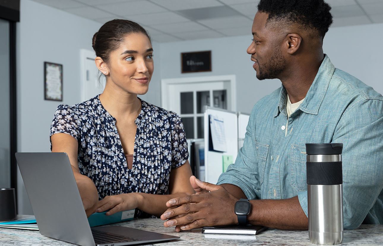 Woman and man sitting at a table, looking at each other and the woman is pointing at a laptop screen