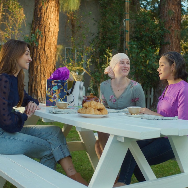 Three women sit at a picnic table discussing cancer and life insurance.