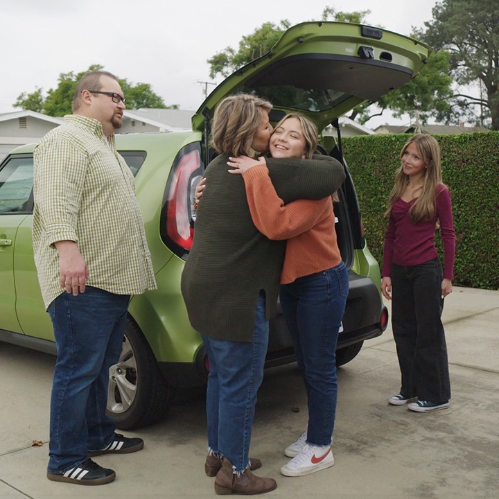 A teenage girl hugs her family in front of her car before leaving for college.